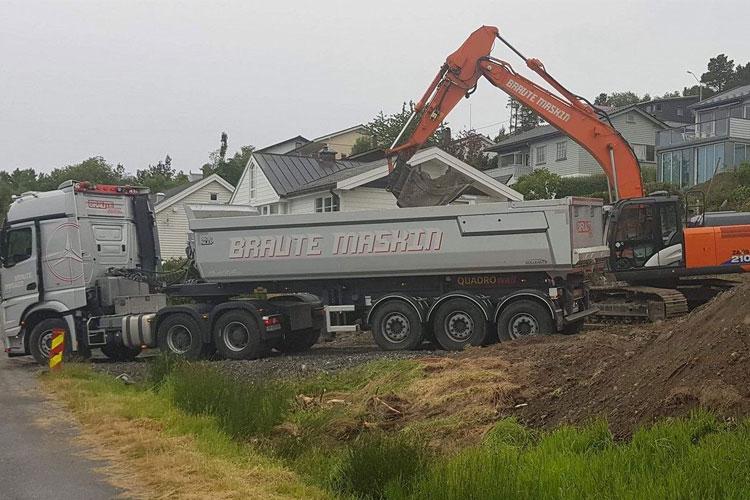 Lastebil med tilhenger og gravemaskin på byggeplass, med hus i bakgrunnen og grønt landskap.
