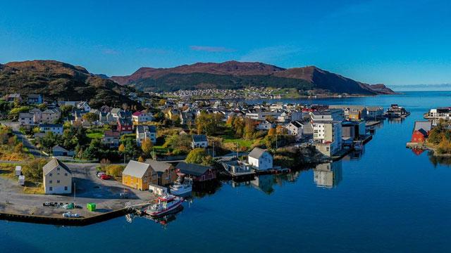 En vakker kystby med fargerike hus, båter i havnen og fjell i bakgrunnen under en klar blå himmel.