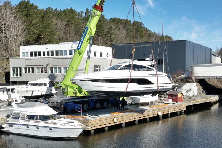 En stor yacht heises opp av en kran ved en båtverft, med flere båter og bygninger i bakgrunnen.