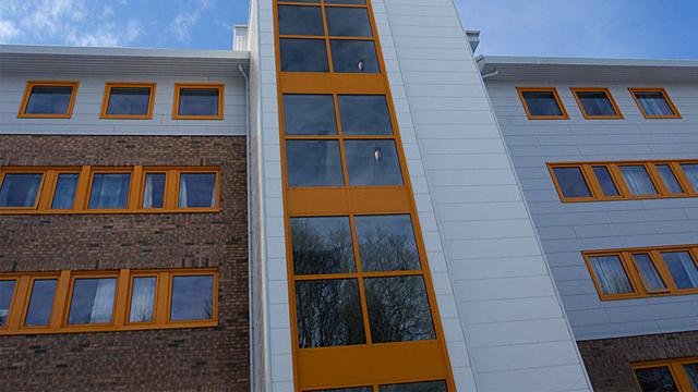 Moderne bygning med store vinduer og oransje detaljer, beliggende under en blå himmel.
