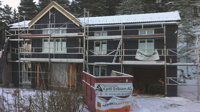 Nybygget hus med stillas og snø på bakken, med en rød container foran.