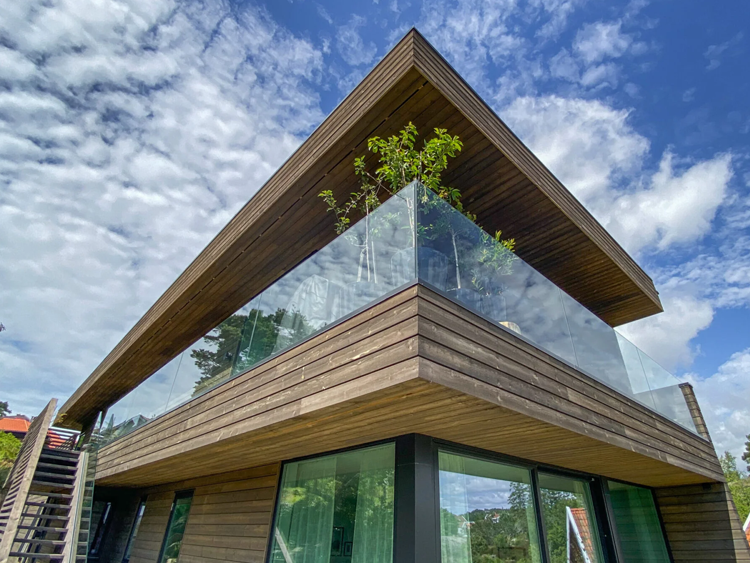 Moderne hus med trepanel og glassbalkong under en blå himmel med skyer.