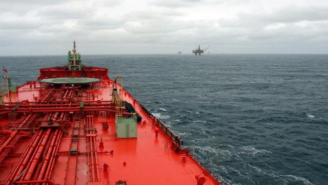 Utsikt fra et rødt tankskip på havet, med en oljeplattform i bakgrunnen under en overskyet himmel.