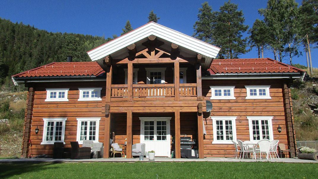 En vakker, moderne hytte i tre med rød takstein, balkong og store vinduer, omgitt av trær og grønt gress.