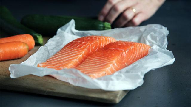 To skiver av laks på papir, med gulrøtter og squash i bakgrunnen på et skjærebrett.