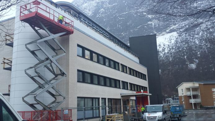 Bygning under oppussing med stillas og arbeidere i fjellandskap. Snø på bakken og grå himmel.