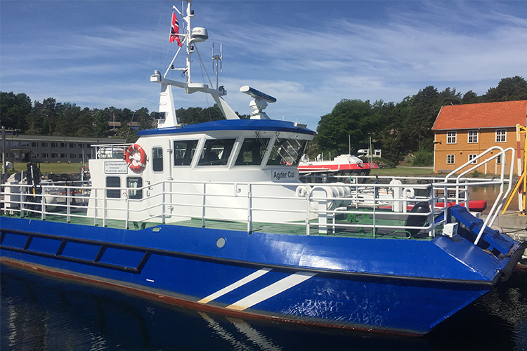 Blå båt med norsk flagg ved kai, omgitt av bygninger og båter i en solfylt havn.