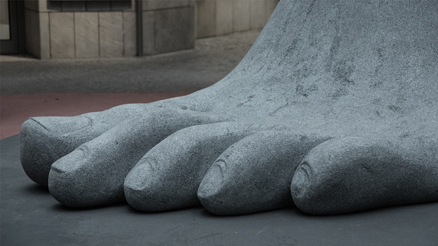 En stor, grå skulptur av en fot med detaljerte tær, plassert på en urban bakgrunn.