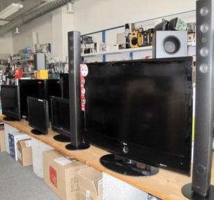 Flate skjermer og høyttalere utstilt i en elektronikkbutikk med diverse tilbehør i bakgrunnen.