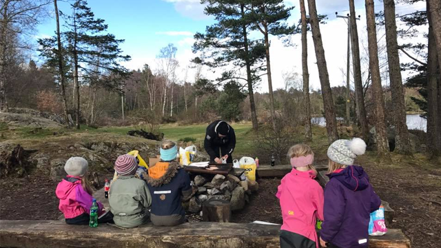 Barn som sitter rundt et bålsted i skogen, mens en voksen lager mat på en benk.