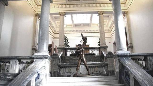 En bronse statue av en mann som hever en arm, plassert i en stor, lys hall med søyler.