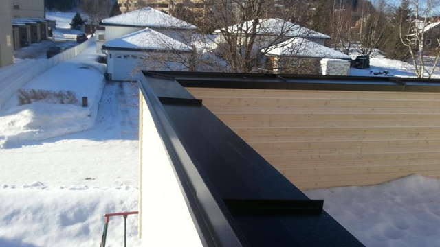 Utsikt fra taket med snø på bakken og hus i bakgrunnen, med en svart takrenne i forgrunnen.