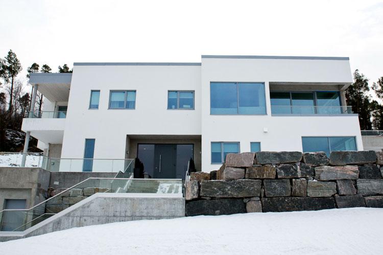 Moderne hvit bolig med store vinduer, beliggende på snødekt område med steinmur foran.