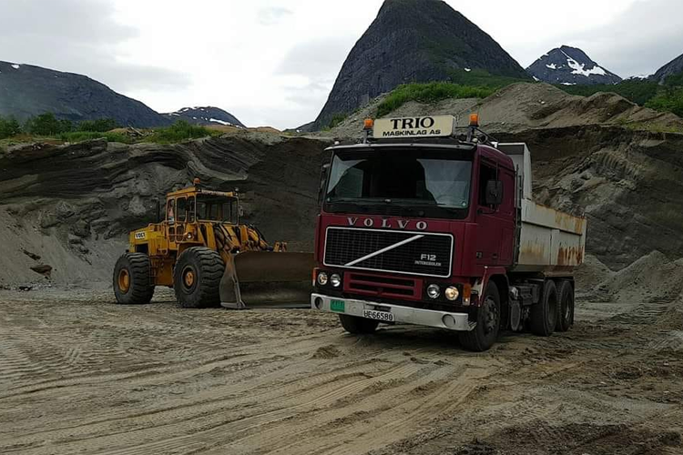 En rød Volvo lastebil og en gult gravemaskin på en byggeplass med fjell i bakgrunnen.