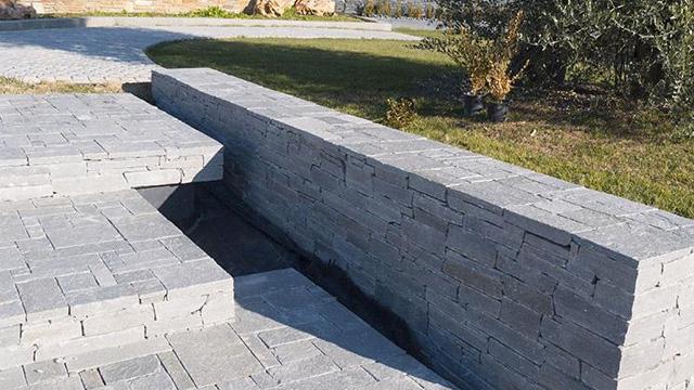 Grå steinmur med moderne design, plassert i en hage med grønt gress og trær i bakgrunnen.