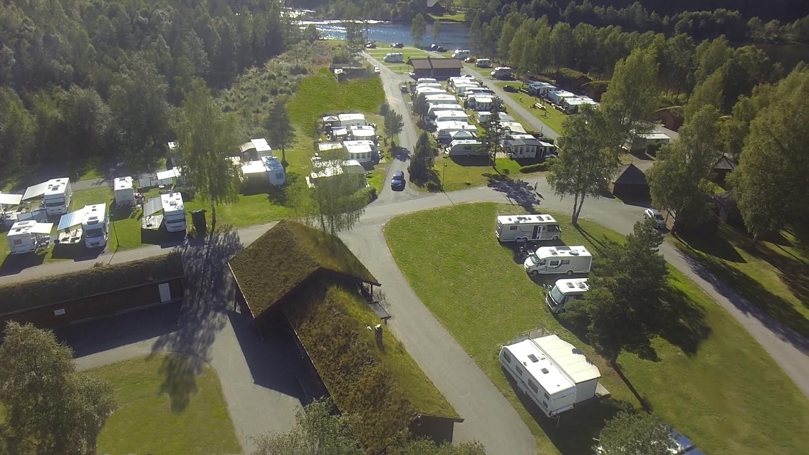 Oversiktsbilde av en campingplass med bobiler, trær og en innsjø i bakgrunnen.