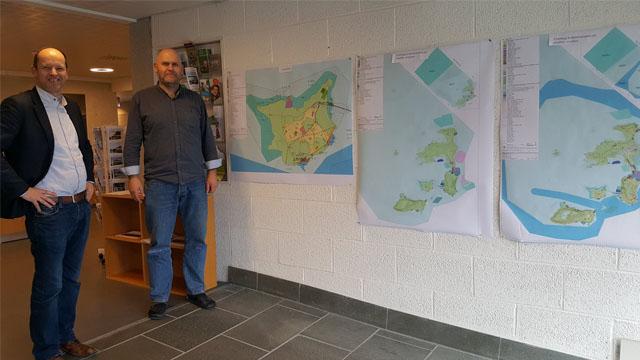 To menn står ved en vegg med kart som viser øyer og områder, i et kontorlandskap.