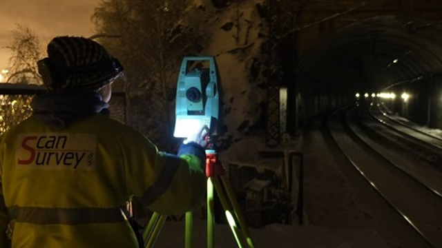 En person i refleksjakke bruker en totalstasjon for å måle i nærheten av en jernbanetunnel om natten.