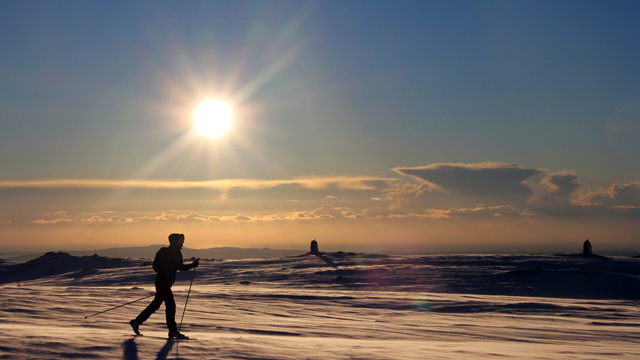 En skiløper går over snødekte fjell under en solfylt himmel med skyer i bakgrunnen.