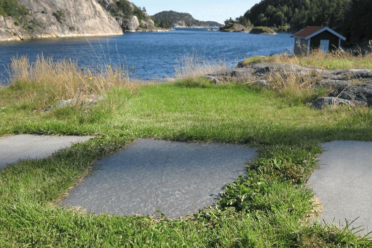Utsikt over en innsjø med steinplater og grønt gress i forgrunnen, omkranset av fjell og trær.
