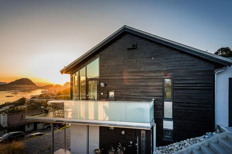 Moderne hus med svart trepanel og glassbalkong, beliggende ved sjøen under solnedgang.