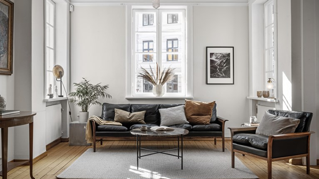Moderne stue med svart sofa, glassbord, planter og store vinduer som slipper inn naturlig lys.