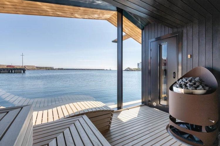 Utsikt fra en moderne terrasse ved vannet med en peis og solfylt himmel.