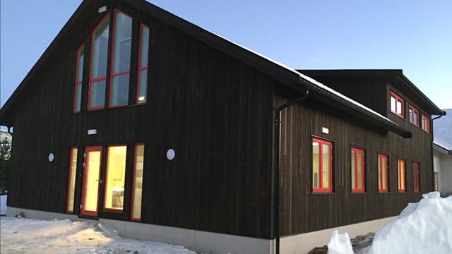 Moderne svart hus med røde vinduer og snø foran, beliggende i et vinterlandskap.