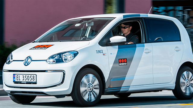 En hvit Volkswagen bil kjører på veien med en sjåfør iført solbriller og logoer på siden.