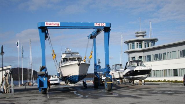 Båter heises opp av en kran ved en marina, med bygninger og blå himmel i bakgrunnen.