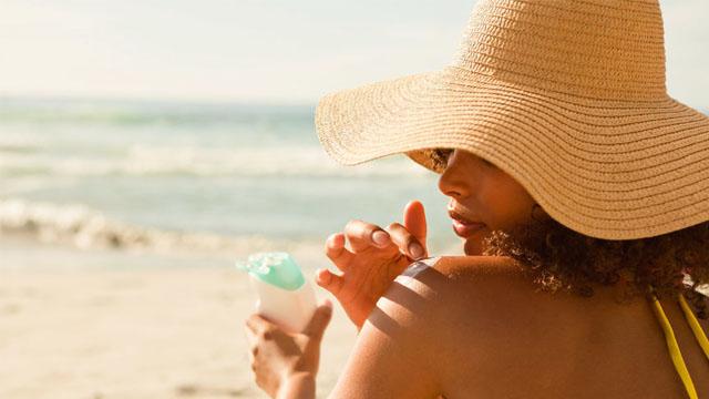 Kvinne med stor solhatt som smører solkrem på armen mens hun nyter is ved stranden.
