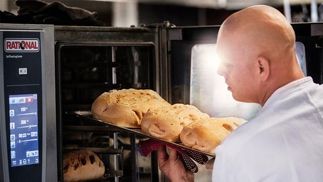 Baker tar ut nybakt brød fra ovnen med en stekeplate i hånden.