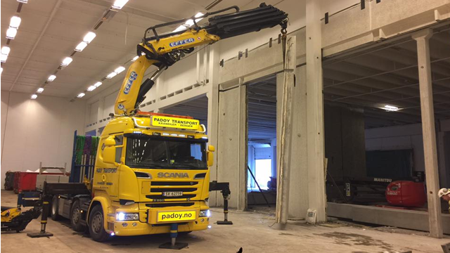 En gul Scania lastebil med kran som løfter materialer i en stor lagerbygning.
