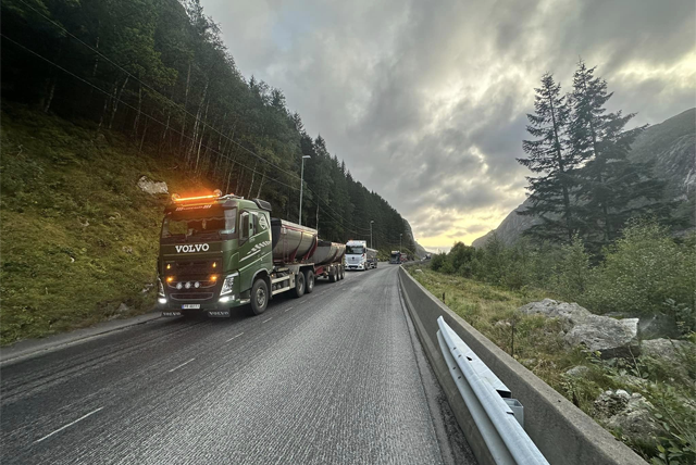 En lastebil fra Volvo kjører på en vei omringet av trær og fjell under en skyet himmel.