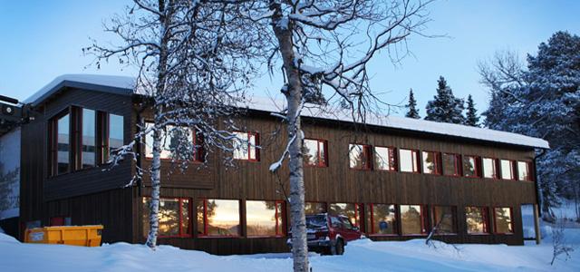 Moderne hytte med store vinduer, omgitt av snø og trær, i en vinterlig fjellandskap.
