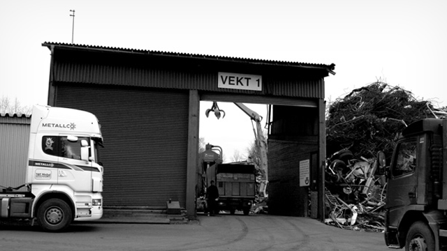 Lastebiler ved inngangen til en resirkuleringsfabrikk med metallskrap i bakgrunnen.