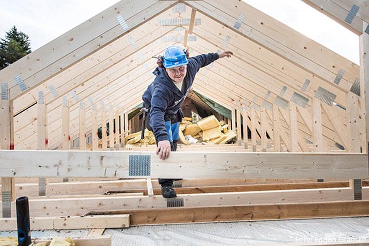 En håndverker i hjelm balanserer på en takstol under bygging av et hus. Byggematerialer er synlige i bakgrunnen.