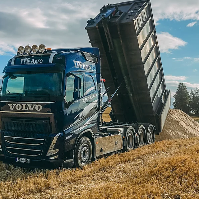 En blå Volvo lastebil tømmer sand i et åkerlandskap under en klar himmel.
