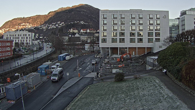 Bygget i bakgrunnen er et hotell, med byggearbeid og fjell i bakgrunnen, samt biler og containere på veien.