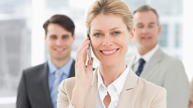 En kvinne i beige dress smiler mens hun snakker i telefonen, med to menn i bakgrunnen.