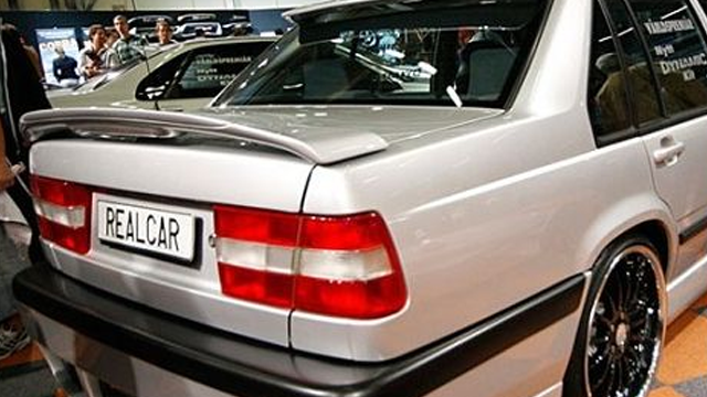 En sølvfarget bil med spoiler, sett fra bak, på en bilutstilling med mennesker i bakgrunnen.