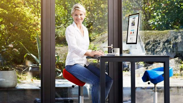 Kvinne som sitter ved et moderne skrivebord med datamaskin, omgitt av grønne planter og naturlig lys.
