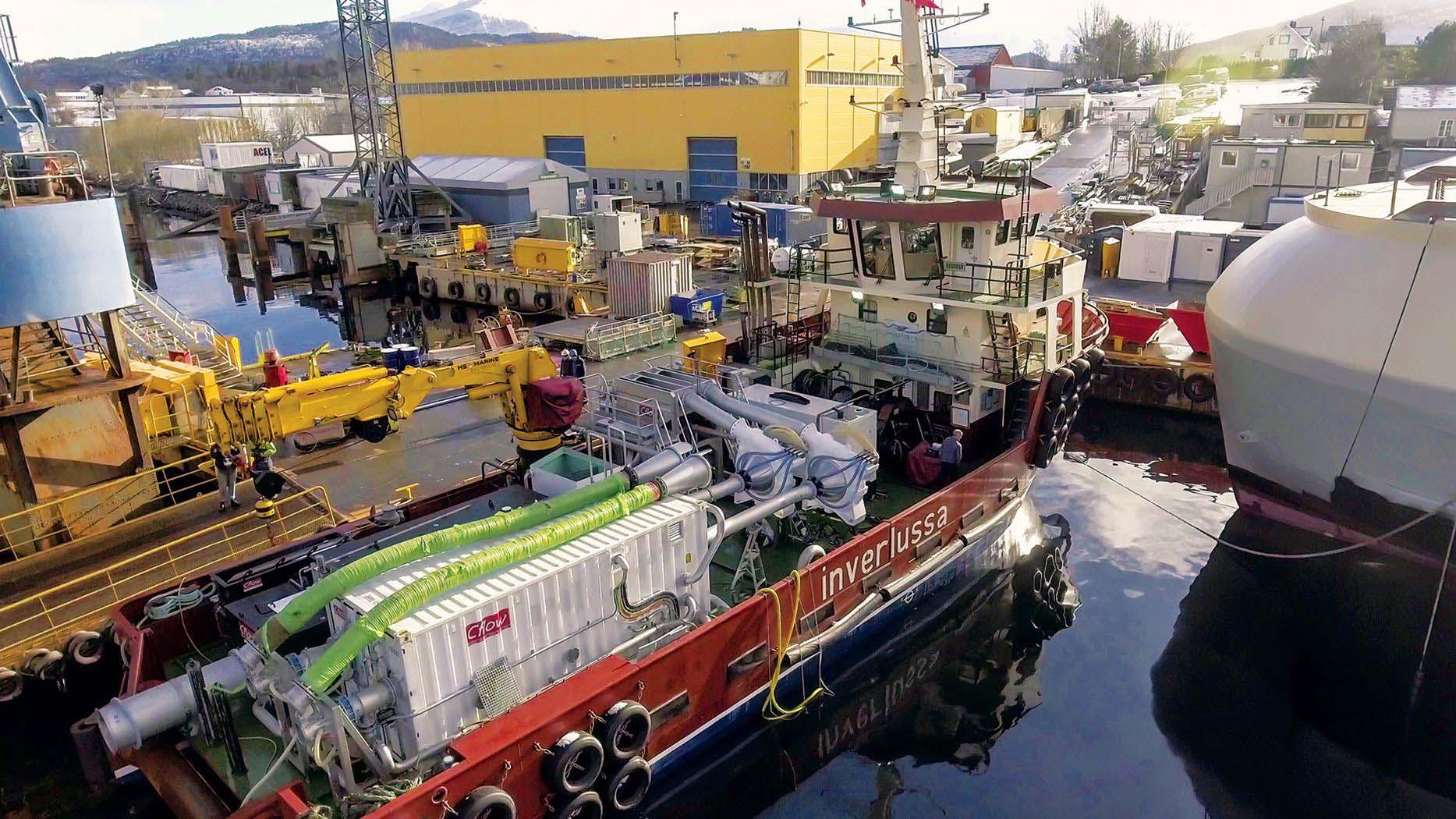 Tugbåt ligger ved kai i havnen, omgitt av ulike båter og industriell infrastruktur. Sollys reflekteres i vannet.
