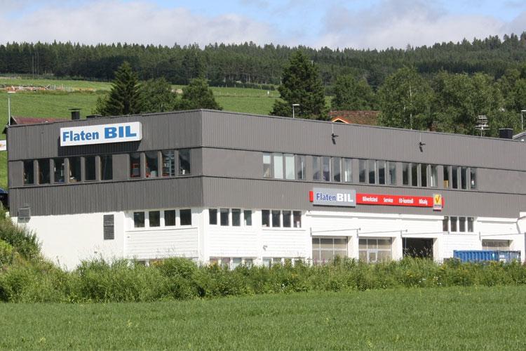Flaten BIL bygning med flere vinduer og logoer, omgitt av grønt landskap og trær i bakgrunnen.