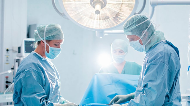 Kirurgisk team i operasjonsstue, iført munnbind og sterile klær, arbeider med pasienten under operasjon.
