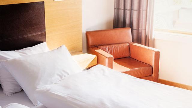 Lys hotellrom med en seng med hvite sengetøy og en brun lenestol ved vinduet.