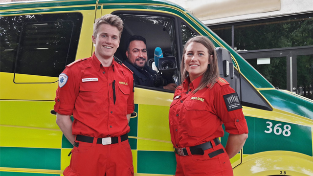 To personer i røde ambulanseuniformer står foran en ambulanse, med en sjåfør i førersetet.