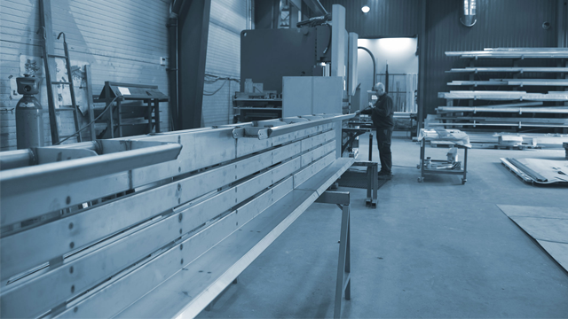 En person jobber med lange metallplater i en industriell verksted med maskiner og utstyr i bakgrunnen.