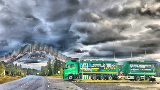 Grønn lastebil med tilhenger står på veien under en dramatisk skyhimmel med en stor kunstinstallasjon i bakgrunnen.