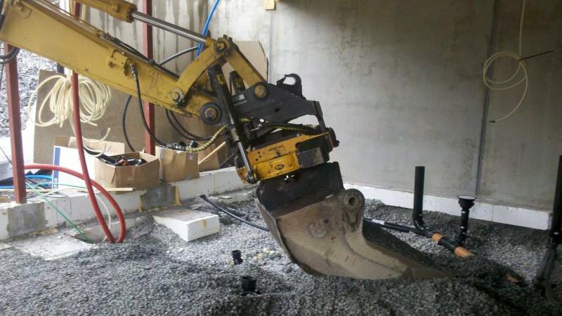Gravemaskin med skuffe som arbeider på byggeplass, med grus og betongfundament i bakgrunnen.